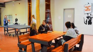 Varian opiskelijoita aulassa istumassa pöydän ääressä ja taustalla kaksi opiskelijaa pelaamassa pingistä.