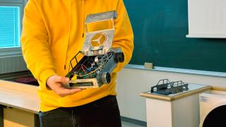 Opiskelija esittelee valmista pienoisautoa, jonka katto on nostettu ylös. Taustalla olevalla pöydällä on toisen auton runko.