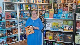 Leena Anttila seisoo isojen lautapelihyllyjen edessä hymyillen ja kädessään Smart10-peli.