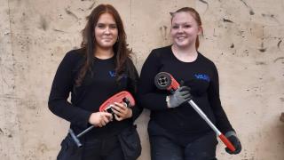 Yolanda ja Leila hymyilevät työvaatteissa työkalut käsissään.