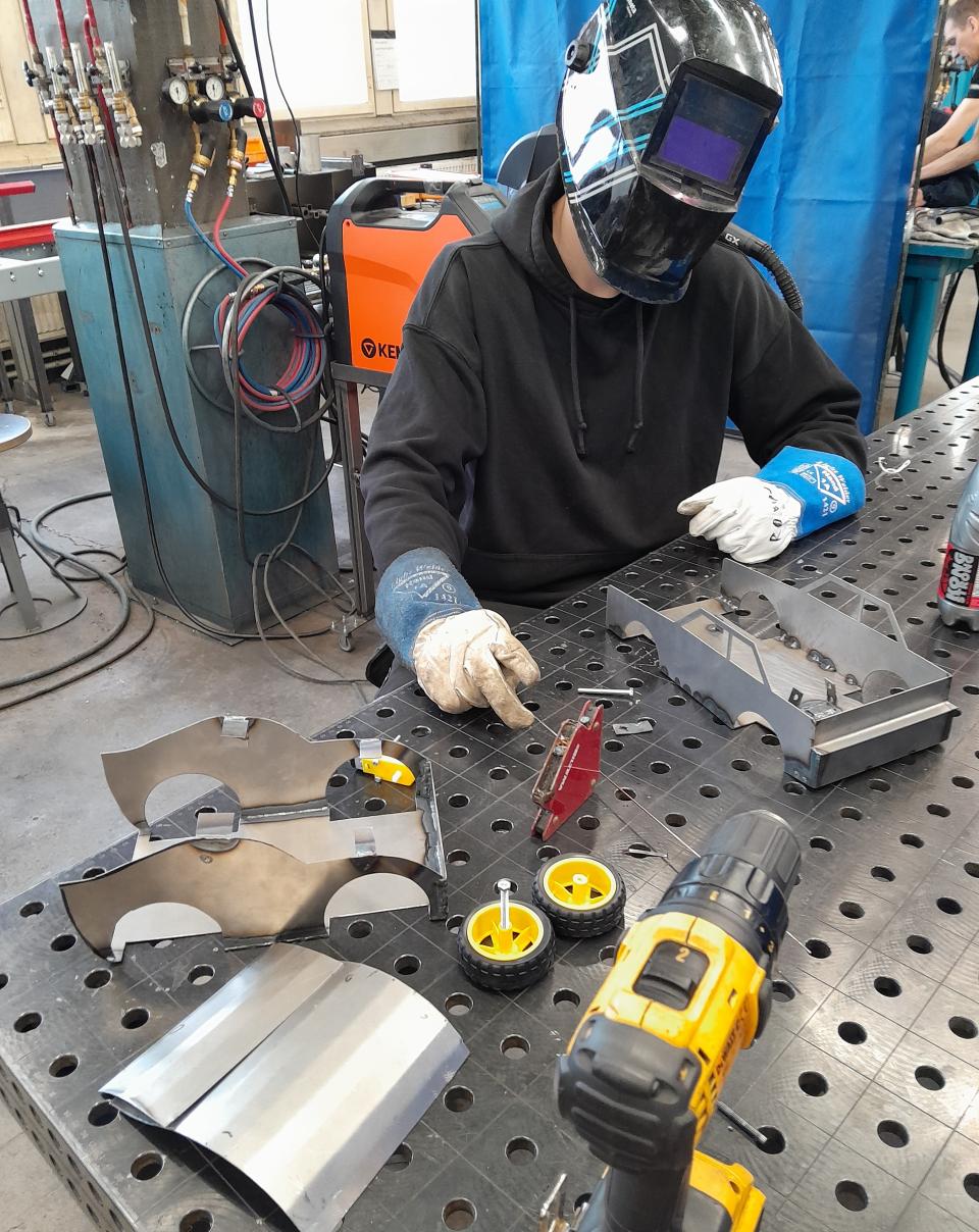 Opiskelija suojakypärässä ja suojahanskoissa työpöydän ääressä.